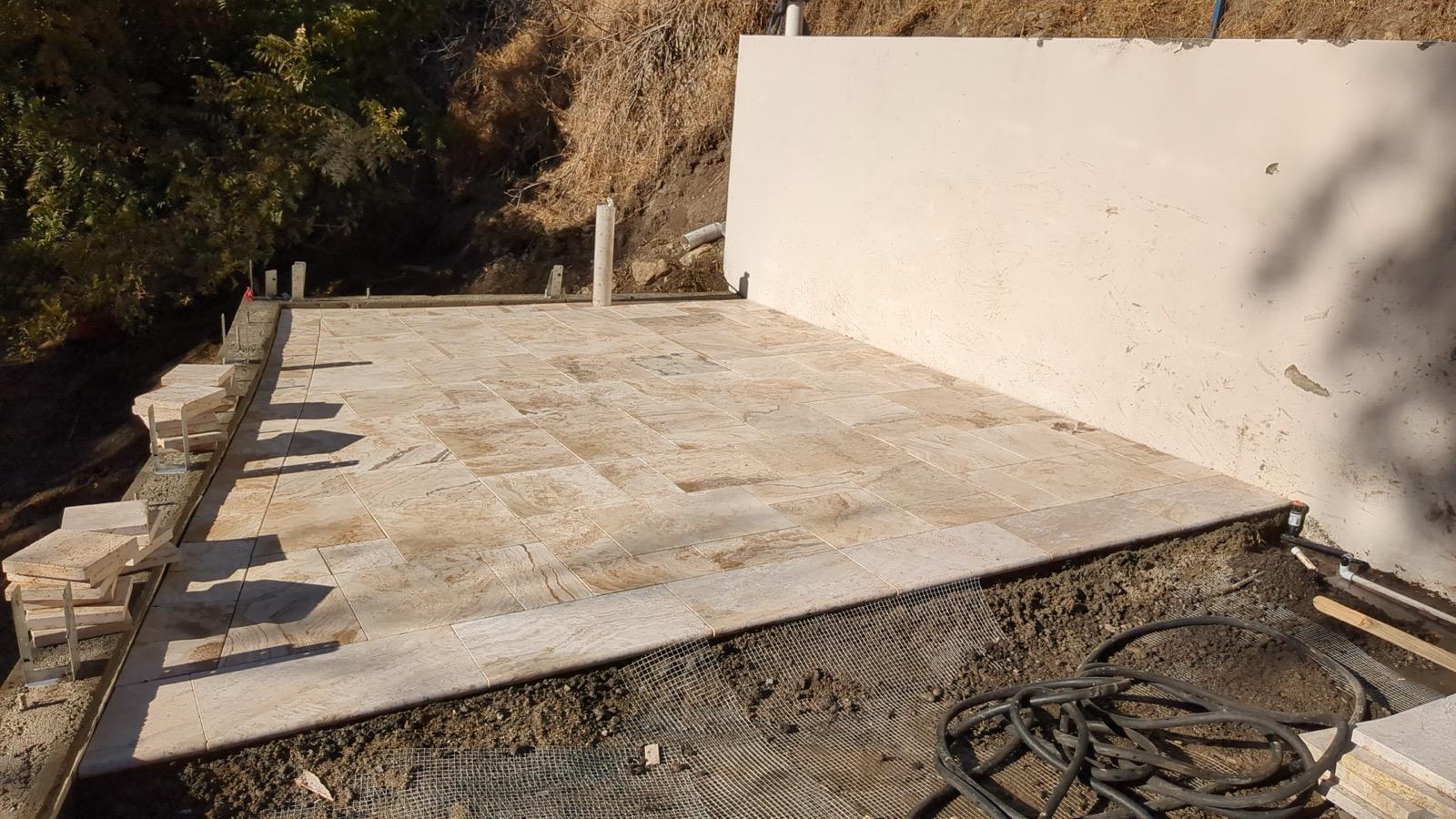 Beige paver patio under construction on a hillside lot, with base prep and irrigation lines in place
