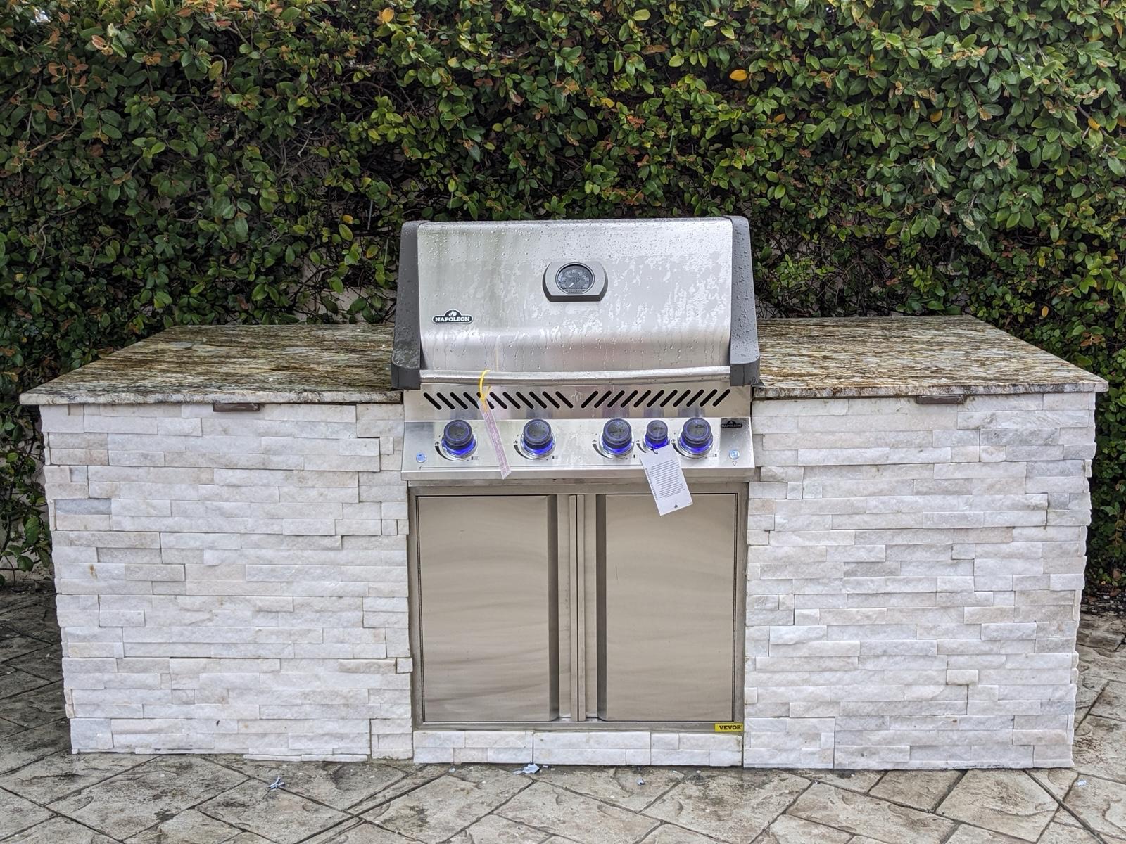 Freestanding outdoor grill island faced in light stacked stone in front of a hedge